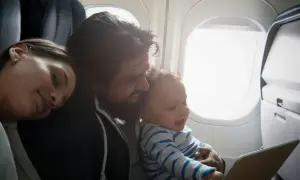 En familj njuter av att flyga med barn. Pappan håller sitt leende barn som tittar på en surfplatta, medan mamman vilar sitt huvud mot hans axel.