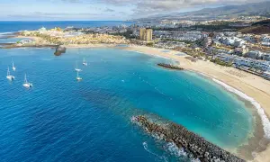 Vacker vy över en strand med turkost vatten och segelbåtar. En stad breder ut sig längs kusten, perfekt för en resa till Teneriffa i november.