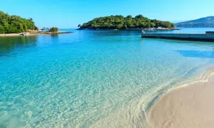 Kristallklart turkost vatten möter en sandstrand vid ett vackert albanien resmål. En grönskande ö syns i bakgrunden.