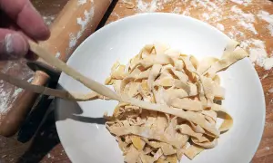 Färska tagliatelle av pastadeg durumvete läggs i en skål. En kavel och mjöl syns i bakgrunden.