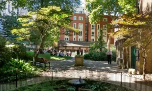 En grönskande park i London med människor som kopplar av på gräsmattan. En fågel sitter på en fontän i förgrunden.