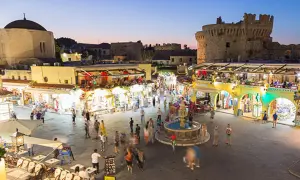 Kvällsliv på Rhodos med folk som njuter av Rhodos shopping och restauranger vid en fontän, med en borg i bakgrunden.