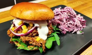 En saftig pulled pork-burgare med majs, rödlök och krämig sås, serverad med en lila kålsallad.