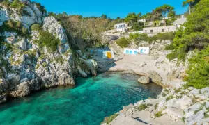 En klippig vik med turkost vatten i Salento, Italien. Vita byggnader med blå dörrar och en gul vägg klättrar uppför sluttningen.