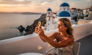 En kvinna tar en selfie med sin telefon på en balkong med utsikt över Santorini. Solnedgången färgar himlen och havet gyllene, perfekt för att dokumentera sitt **båtluffa Grekland** äventyr.