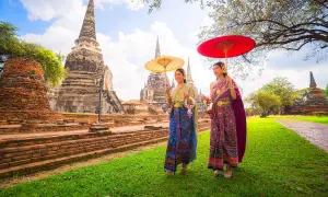 Två kvinnor i traditionella thailändska kläder med parasoller promenerar vid gamla tempelruiner, en scen som påminner om den kultur som talas i Bangkok.