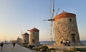 Gamla väderkvarnar längs kajen i Rhodos stad. Folk promenerar och njuter av kvällssolen vid havet.
