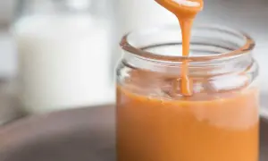 A spoon pours smooth, glossy caramel sauce into a glass jar.