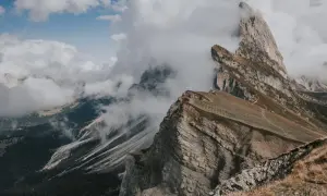 Molnen sveper in över dramatiska bergstoppar i Dolomiterna, Italien. En vandringsled slingrar sig uppför en gräsklädd sluttning.