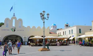 Solig dag på **Kos stad**s torg. Folk sitter vid caféer under parasoller framför en gul byggnad med grekisk flagga. Blå kupoler syns i bakgrunden.