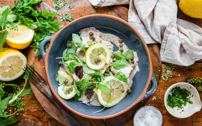 Vitello tonnato i en skål, toppad med citronskivor, kapris och ruccola. Serveras med citroner och färska örter.