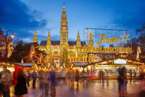 En livlig julmarknad i Europa med folk som strosar bland ljus och stånd framför en vackert upplyst byggnad.