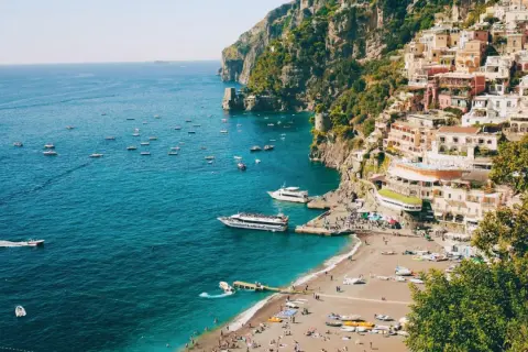 Färgstark by klättrar uppför klipporna vid sorrentohalvön, med båtar på det turkosa havet och en strand full av folk.