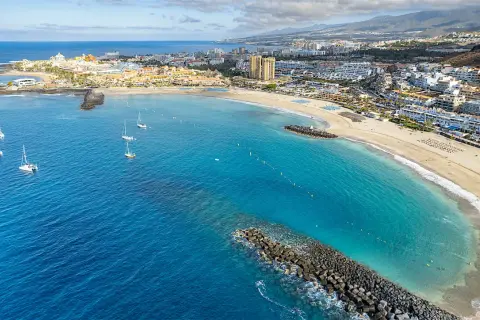 Vacker vy över en strand med turkost vatten och segelbåtar. En stad breder ut sig längs kusten, perfekt för en resa till Teneriffa i november.