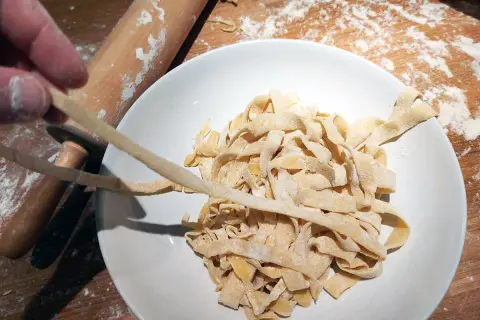 Färska tagliatelle av pastadeg durumvete läggs i en skål. En kavel och mjöl syns i bakgrunden.