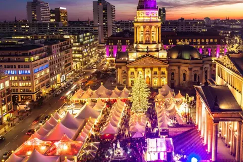 En livlig julmarknad i Berlin med tält, en stor julgran och upplysta byggnader i skymningen.