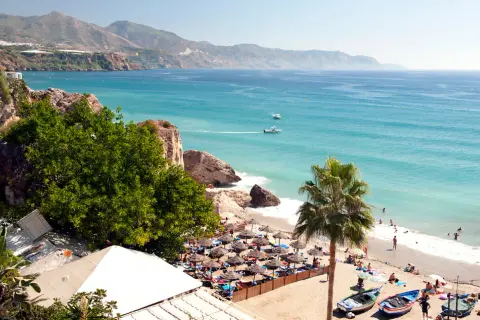 En solig strand med parasoller och badande människor. Vackert turkost hav och berg i bakgrunden. Spanien temperatur året runt är perfekt för detta.