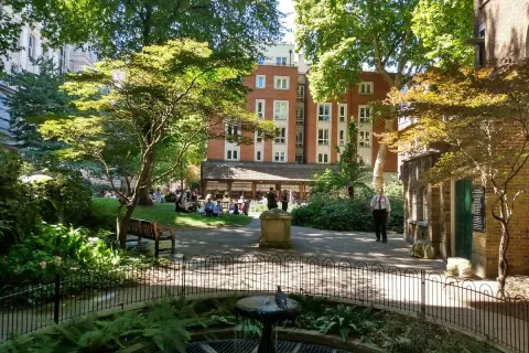 En grönskande park i London med människor som kopplar av på gräsmattan. En fågel sitter på en fontän i förgrunden.