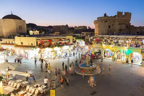 Kvällsliv på Rhodos med folk som njuter av Rhodos shopping och restauranger vid en fontän, med en borg i bakgrunden.