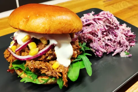 En saftig pulled pork-burgare med majs, rödlök och krämig sås, serverad med en lila kålsallad.