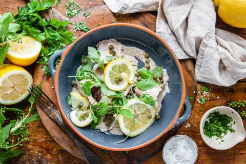 Vitello tonnato i en skål, toppad med citronskivor, kapris och ruccola. Serveras med citroner och färska örter.
