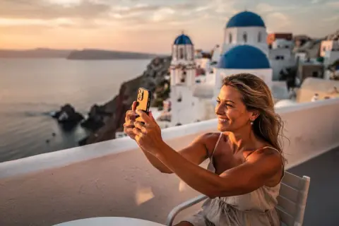 En kvinna tar en selfie med sin telefon på en balkong med utsikt över Santorini. Solnedgången färgar himlen och havet gyllene, perfekt för att dokumentera sitt **båtluffa Grekland** äventyr.