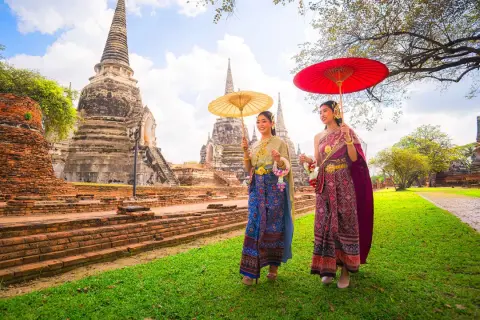 Två kvinnor i traditionella thailändska kläder med parasoller promenerar vid gamla tempelruiner, en scen som påminner om den kultur som talas i Bangkok.