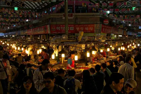 Livligt folkliv på Gwangjang Market, med matstånd som säljer koreansk mat under blinkande lampor och flaggor.