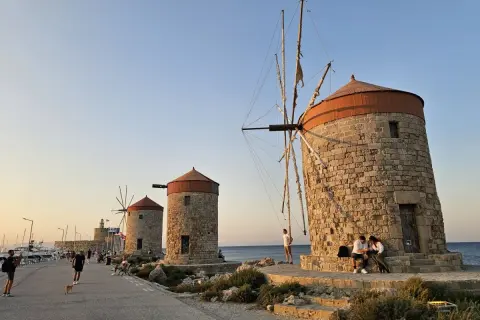 Gamla väderkvarnar längs kajen i Rhodos stad. Folk promenerar och njuter av kvällssolen vid havet.