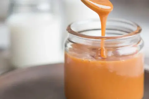 A spoon pours smooth, glossy caramel sauce into a glass jar.
