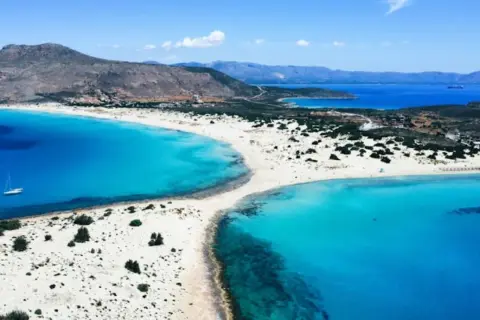 En vit sandstrand med turkost vatten och en segelbåt vid **Platanias, Kreta**. Berg i bakgrunden.