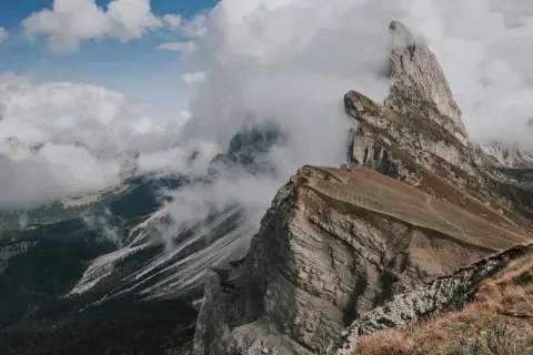 Molnen sveper in över dramatiska bergstoppar i Dolomiterna, Italien. En vandringsled slingrar sig uppför en gräsklädd sluttning.