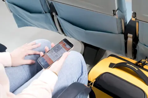 Person visar boardingkort på mobilen i ett flygplanssäte. Gul Samsonite-väska bredvid. Smidigt med digitala biljetter för ditt ving flygbolag.