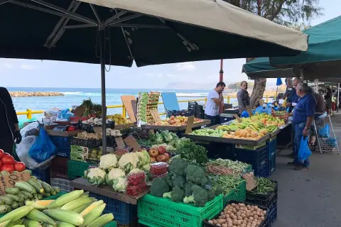 Frukt- och grönsaksstånd vid marknad i Chania. Färska varor som majs, broccoli och tomater säljs med havet i bakgrunden.