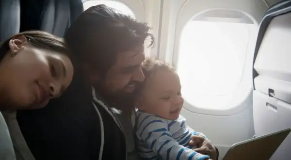 En familj njuter av att flyga med barn. Pappan håller sitt leende barn som tittar på en surfplatta, medan mamman vilar sitt huvud mot hans axel.