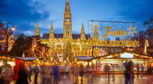 En livlig julmarknad i Europa med folk som strosar bland ljus och stånd framför en vackert upplyst byggnad.