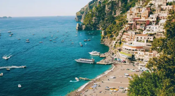 Färgstark by klättrar uppför klipporna vid sorrentohalvön, med båtar på det turkosa havet och en strand full av folk.