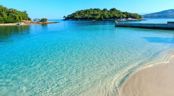Kristallklart turkost vatten möter en sandstrand vid ett vackert albanien resmål. En grönskande ö syns i bakgrunden.