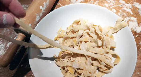 Färska tagliatelle av pastadeg durumvete läggs i en skål. En kavel och mjöl syns i bakgrunden.