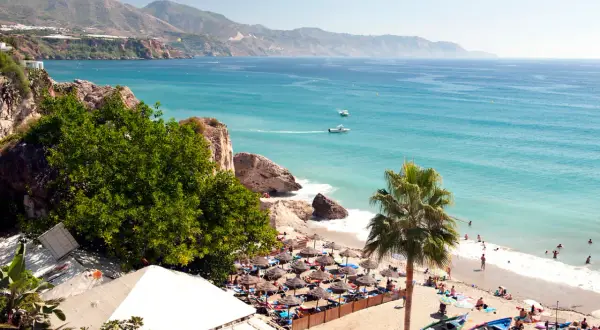 En solig strand med parasoller och badande människor. Vackert turkost hav och berg i bakgrunden. Spanien temperatur året runt är perfekt för detta.