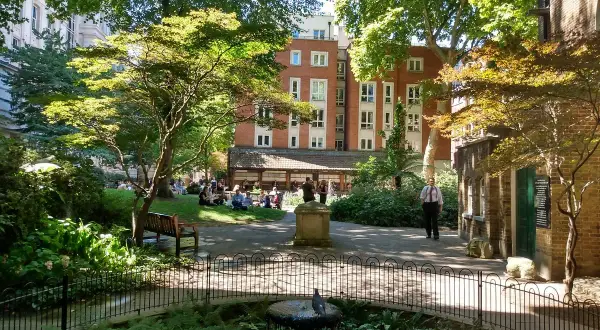 En grönskande park i London med människor som kopplar av på gräsmattan. En fågel sitter på en fontän i förgrunden.
