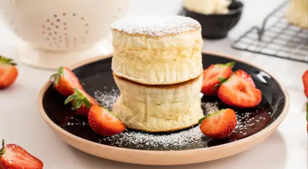 Två fluffiga japanska pannkakor staplade på en tallrik med färska jordgubbar och pudersocker.