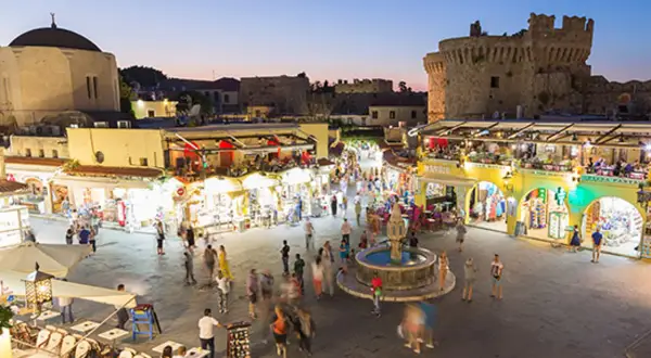 Kvällsliv på Rhodos med folk som njuter av Rhodos shopping och restauranger vid en fontän, med en borg i bakgrunden.