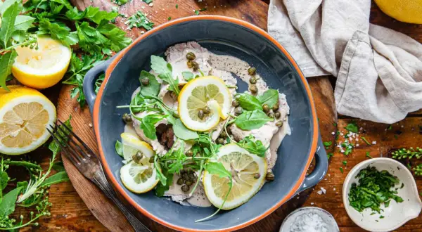 Vitello tonnato i en skål, toppad med citronskivor, kapris och ruccola. Serveras med citroner och färska örter.