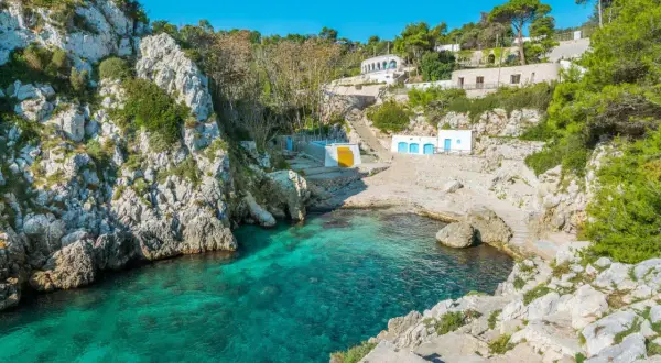 En klippig vik med turkost vatten i Salento, Italien. Vita byggnader med blå dörrar och en gul vägg klättrar uppför sluttningen.