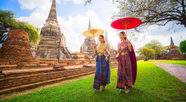 Två kvinnor i traditionella thailändska kläder med parasoller promenerar vid gamla tempelruiner, en scen som påminner om den kultur som talas i Bangkok.