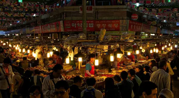 Livligt folkliv på Gwangjang Market, med matstånd som säljer koreansk mat under blinkande lampor och flaggor.
