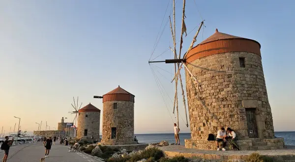 Gamla väderkvarnar längs kajen i Rhodos stad. Folk promenerar och njuter av kvällssolen vid havet.
