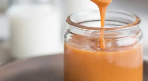 A spoon pours smooth, glossy caramel sauce into a glass jar.
