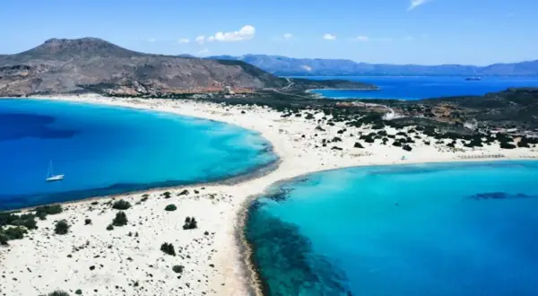 En vit sandstrand med turkost vatten och en segelbåt vid **Platanias, Kreta**. Berg i bakgrunden.
